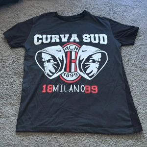 Svart AC Milan Curva Sud t-shirt XS - Svart t-shirt med tryck framtill: 'CURVA SUD', två ansikten med huvor och AC Milan-logga samt texten '18 MILANO 99' i vitt och rött. Klassisk rund hals och korta ärmar. Perfekt för dig som älskar fotboll och italiensk stil.