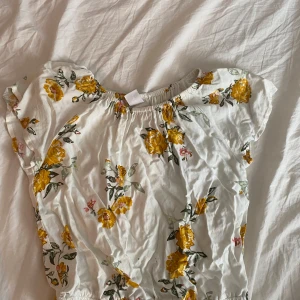 Vit blommig blus med gula rosor - Supersöt vit blus med gula och rosa blommor, elastisk halsringning och midja för en avslappnad och luftig känsla. Korta ärmar med lätt volang och romantiskt blommönster gör den perfekt för vår och sommar. Materialet känns lätt och andas bra. Tröjan är en avklippt byxdress de är därför de vid midjan som man ser på bild 3 men de går att klippa bort utan problem. Pris kan diskuteras 
