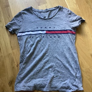 Grå t-shirt Tommy Hilfiger XS - Snygg grå t-shirt från Tommy Hilfiger i bomull. Klassisk rund halsringning och korta ärmar. Framsidan har det ikoniska Hilfiger-trycket med röd, vit och marinblå rand. Perfekt basic med en clean vibe och skön passform.