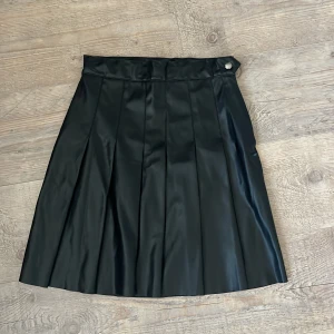 Svart plisserad kjol i skinnlook SRNHILT - Svart plisserad kjol från SRNHILT i skinnimitation med snygg midja, dragkedja i sidan och knappdetalj. Kjolen har en cool, glansig finish och klassisk A-linjeform som ger en edgy vibe. Perfekt för dig som vill sticka ut med en stilren look.