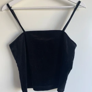 Svart sammetstopp med smala axelband - En stilren svart topp i mjuk sammet med smala axelband och rak halsringning. Perfekt för dig som gillar minimalistisk och trendig stil. Toppen är croppad och har en enkel, elegant look som passar till många outfits.