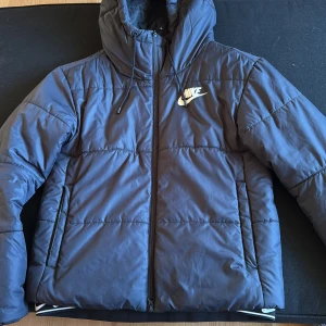 Blå pufferjacka från Nike med huva - Säljer en blå pufferjacka från Nike med huva och dragsko. Jackan har dragkedja framtill, två sidofickor och klassisk Nike-logga på bröstet. Perfekt för kalla dagar när du vill hålla stilen och vara varm.