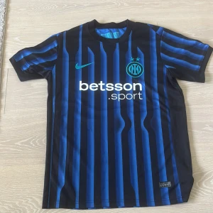 Inter Milan randig fotbollströja Nike - Säljer en officiell Inter Milan fotbollströja från Nike med blå och svarta vertikala ränder, klubbmärke och sponsortryck på bröstet. Tröjan har korta ärmar, rund hals och är gjord i lätt, ventilerande polyester. UEFA Foundation patch på ärmen.