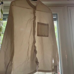 Beige sidenliknande skjorta med ficka - Snygg beige skjorta med lång ärm och rund krage. Materialet är tunt och har en sidenliknande glans. Skjortan har en bröstficka och knappar hela vägen framtill. Perfekt för dig som gillar minimalistisk och stilren design.