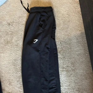 Svarta träningsbyxor från Gymshark - Svarta träningsbyxor från Gymshark med elastisk midja och snörning. Byxorna har en stilren design med Gymshark-logga på benet och är gjorda i ett lätt syntetmaterial som passar perfekt för gymmet eller andra sportaktiviteter.