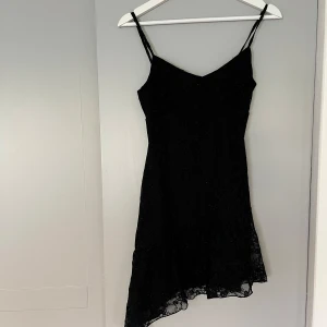 Svart spetsklänning med asymmetrisk volang - Supersnygg svart kortklänning med tunna axelband och v-ringad hals. Klänningen har ett lager av glittrig spets med blommigt mönster och en asymmetrisk volangkant nedtill som ger en cool och unik look. Perfekt för dig som gillar detaljer och vill sticka ut.