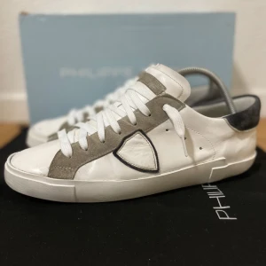 Philippe Model sneakers  - Snygga vita sneakers från Philippe Model med grå mockadetaljer och vit snörning. Skorna har en rund tå, platt sula och en cool sköldlogga på sidan. Bakkappan är i mörkgrå mocka och ovandelen är i skinn. Perfekta för dig som gillar stilrena och trendiga sneakers. Storlek 42, bra skick, endast skor medföljer! Sulan är reparerad av skomakare! Nypris runt 3500 kr! Hör av er ifall ni har några frågor! 🙌⚜️