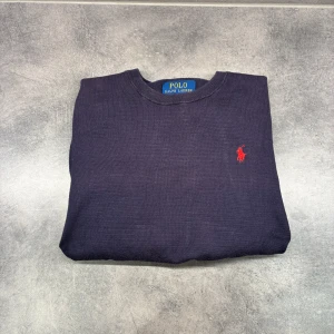 Marinblå polo tröja  - Snygg mörkblå stickad tröja från Polo Ralph Lauren med klassisk rund hals och röd broderad logga på bröstet. Tröjan har långärmad passform och är gjord i mjukt material som sitter skönt. Perfekt för dig som gillar stilrena och tidlösa plagg.