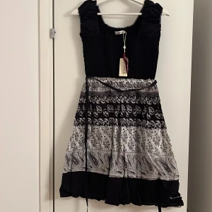 Svart och vit kortklänning från  bondelid  - Supersöt kortklänning från Bracke med svart smockad överdel och breda axelband. Kjolen har ett unikt mönster i svart och vitt med paisley och medaljongdetaljer, samt ett smalt knytband i midjan. Perfekt för dig som gillar boho-vibe och coola prints.
