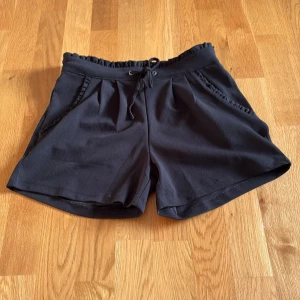 Svarta shorts med snörning från JDY - Svarta shorts från JDY med elastisk midja och snörning framtill. Modellen har två fickor med diskret volangkant och är tillverkad i ett mjukt, lätt material som passar perfekt för varma dagar. Enkel och stilren design som är lätt att matcha.