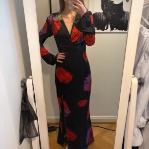Svart långklänning med blommor från Adoore - Supersnygg svart långklänning från Adoore med stora röda och lila blommor. Djup v-ringning, långa ärmar och slits framtill. Aldrig använd pga köpte fel storlek.