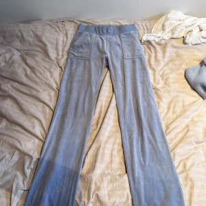 Blå velourbyxor Juicy Couture XS - Snygga ljusblå mjukisbyxor från Juicy Couture i velour med bred resår i midjan, stora fickor fram och bak samt broderad JC-logga på ena fickan. Byxorna har raka ben och klassisk 00-talsvibe. Perfekta för en chill och trendig look. Tvättas innan de skickas 