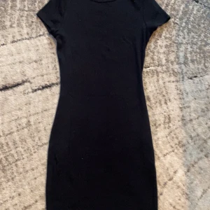 Svart kortklänning från FB Sister - En stilren svart kortklänning från FB Sister i storlek XS. Klänningen har en enkel, figurnära passform med korta ärmar och rund hals. Materialet är mjukt och stretchigt, perfekt för dig som gillar minimalistisk och clean look.