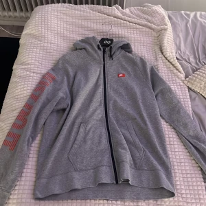 Grå Nike hoodie med röd logga XL - Grå hoodie från Nike i storlek XL med dragkedja och huva. Röd Nike-logga på bröstet och röd text längs ena ärmen. Mjuk bomullsblandning, två fickor framtill och klassisk streetstil. Perfekt för chill dagar eller när du vill ha en sportig look.
