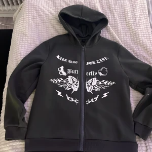 Svart hoodie med tryck från ROMWE - Cool svart hoodie från ROMWE i storlek L med dragkedja och huva. Framsidan har vitt tryck med fjärilar, eld, kedjor och text. Ärmarna har också tryck med taggtråd och fjärilar. Perfekt för dig som gillar streetstyle och unika detaljer.