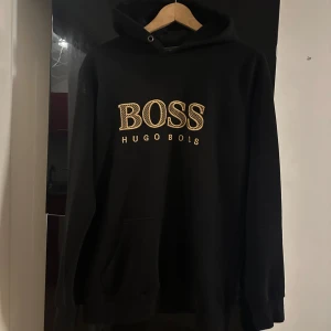 Svart hoodie från Hugo Boss - Svart hoodie från Hugo Boss med stor guldfärgad BOSS-logga och text på bröstet. Tröjan är i storlek S och är inte så använd. Säljs pga den ej passar min stil längre. 