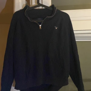 Mörkblå stickad half zip från GANT - Säljer en mörkblå stickad tröja från GANT med half zip-dragkedja och diskret logga på bröstet. Tröjan har ribbad struktur och långa ärmar, perfekt för lager på lager under kyliga dagar.