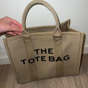 Beige The Tote Bag från Marc Jacobs - Säljer en beige The Tote Bag från Marc Jacobs. Väskan har svarta tryckta bokstäver framtill, två rejäla handtag och en bred avtagbar axelrem. Invändigt finns flera fack och dragkedja för extra förvaring. Har ett stort utrymme och perfekt för datorer och böcker.😊