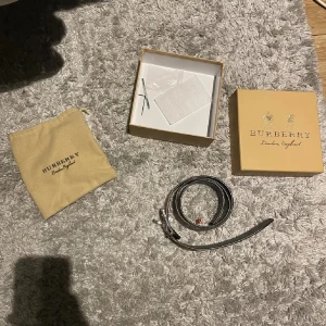Burberry randigt bälte i skinn - Säljer ett snyggt Burberry-bälte i äkta skinn med klassiskt randigt Burberry-mönster i beige, brun och röd. Bältet har en stilren guldfärgad metallspänne och kommer med originalförpackning och dustbag. Perfekt accessoar för att lyfta din outfit.