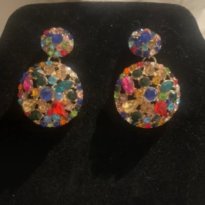 Glittriga örhängen med färgglada stenar - Säljer ett par stora, runda örhängen med massor av glittriga stenar i olika färger som blå, röd, grön, rosa och gul. Örhängena har en cool och festlig vibe med guldtonad bas och är perfekta för att sticka ut. De är täckta av glittrande stenar i olika former och storlekar.