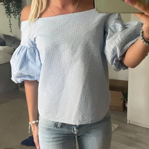 Ljusblå offshoulder blus med puffärm - Superfin ljusblå blus med offshoulder och stora puffärmar. Toppen har en strukturerad, lite krispig yta och är perfekt för dig som gillar en luftig och trendig look. Passar snyggt till jeans eller kjol för en fräsch stil.