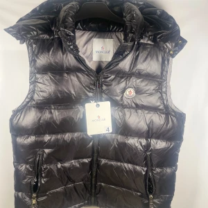 Svart dunväst från Moncler 4/L - Svart dunväst från Moncler med glansig finish och quiltad design. Västen har två fickor med dragkedja, hög krage och klassisk Moncler-logga på bröstet. Perfekt för lager-på-lager och snygg till streetwear. Dragkedja framtill och coolt innerfoder med serietidningsmotiv. Skriv vid frågor storlek 4L
