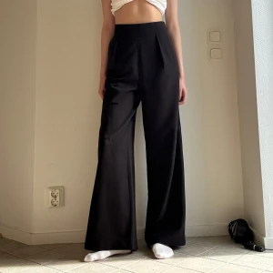 Svarta vida kostymbyxor - Säljer ett par svarta kostymbyxor med hög midja och breda ben. Byxorna har en stilren look med pressveck framtill och sitter snyggt i midjan. Materialet känns mjukt och följsamt. Jag är 173 cm lång.