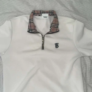 Limited Edition burberry tröja - Snygg vit pikétröja från Burberry med klassisk rutig krage i beige, svart och rött. Tröjan har half zip-dragkedja och broderad Burberry-logga på bröstet. Perfekt för dig som vill ha en clean men ändå lyxig look.