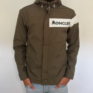 Olivgrön vindjacka från Moncler - Olivgrön vindjacka från Moncler med vit logga på bröstet och huvan. Jackan har huva och vita detaljer på axeln samt Moncler-märke på ärmen. (Jackan är en gammal model som jag fått i arv så därför har jag inget kvitto, den är helt äkta)