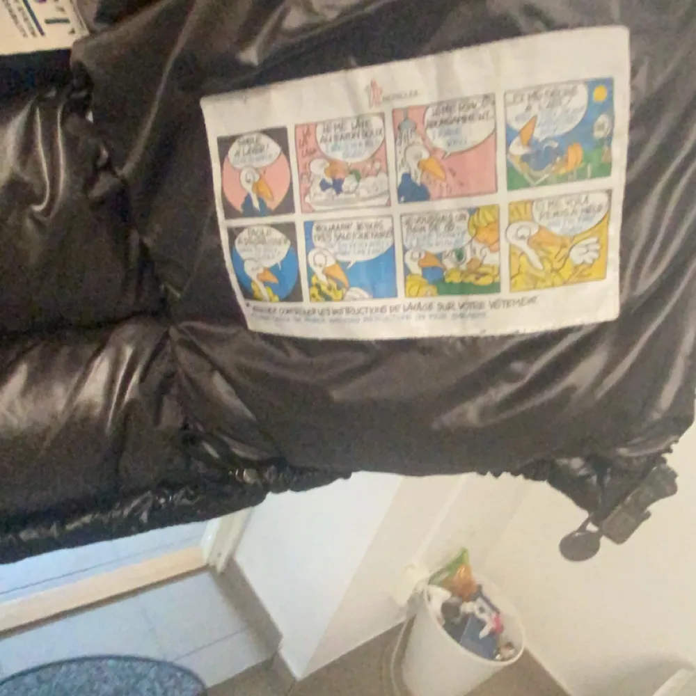 Säljer en svart, glansig pufferjacka från Moncler, moncler maya. med huva och dragkedja. Jackan har lång ärm, två fickor med dragkedja och klassisk Moncler-logga på ärmen. Insidan har ett serietidningsmönster och märkning. Perfekt för kalla dagar.. Takit.