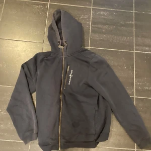 Svart hoodie från Sail Racing - Mörkgrå hoodie från Sail Racing med dragkedja och huva. Jackan har en bröstficka med dragkedja och justerbar snörning i huvan. Perfekt för kyliga dagar och enkel att matcha med det mesta.