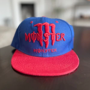 Blå och röd Monster keps - Säljer en blå keps med röd skärm och broderad Monster-logga i rött framtill. Kepsen är justerbar! Synligt använd på sidan och på spännet inuti men inget man direkt ser vid första anblick eller framifrån!