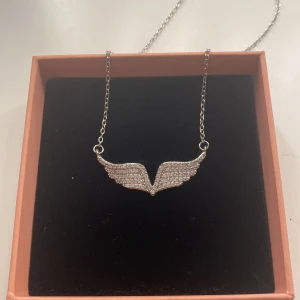 Silver halsband med glittriga ängels vingar  - Säljer ett snyggt halsband som ändå sticker ut lite och piffar upp din outfit!  Hänget är i form av glittriga vingar. Kedjan och hänget är i silverfärg och vingarna är täckta av små gnistrande stenar❣️