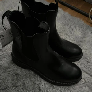 Svarta chelsea boots - Säljer ett par svarta chelsea boots med elastiska paneler på sidorna och dragflikar fram och bak. Skorna har en rund tå och platt sula, perfekta för en stilren look. Helt oanvända och prislappen kvar, tyvärr inte originalförpackningen.