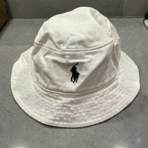 Vit bucket hat från Polo Ralph Lauren - Snygg vit bucket hat från Polo Ralph Lauren med det klassiska broderade logotypen i svart. Perfekt för en stilren look. (Har gulnat lite) storleken är runt 58-59 cm.