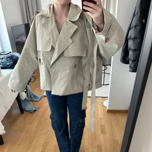 Beige croppad trenchcoat med bälte - Säljer en beige croppad trenchcoat med bälte och stora slag. Jackan har dubbelknäppning med knappar, långa ärmar och en oversized passform. Perfekt för dig som gillar en trendig och avslappnad stil. Säljer för att den är för stor för mig. Passar storleken M