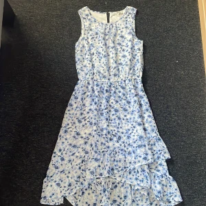 Blommig sommarklänning från Hampton Republic - Säljer en ljusblå och vit ärmlös klänning från Hampton Republic med blommigt mönster. Klänningen har volangdetaljer och en asymmetrisk nederdel som ger en luftig känsla. Perfekt för varma dagar!