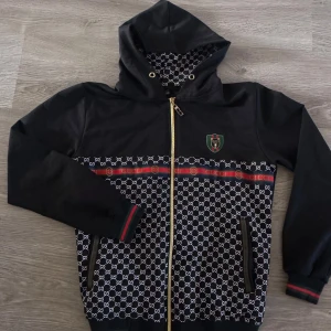 Svart hoodiejacka Gucciliknande med mönster - Svart hoodie med dragkedja, huva och guldiga detaljer. Jackan har ett vitt monogrammönster, broderad logotyp på bröstet och röda/gröna ränder. Fickor med dragkedja och ribbade muddar.