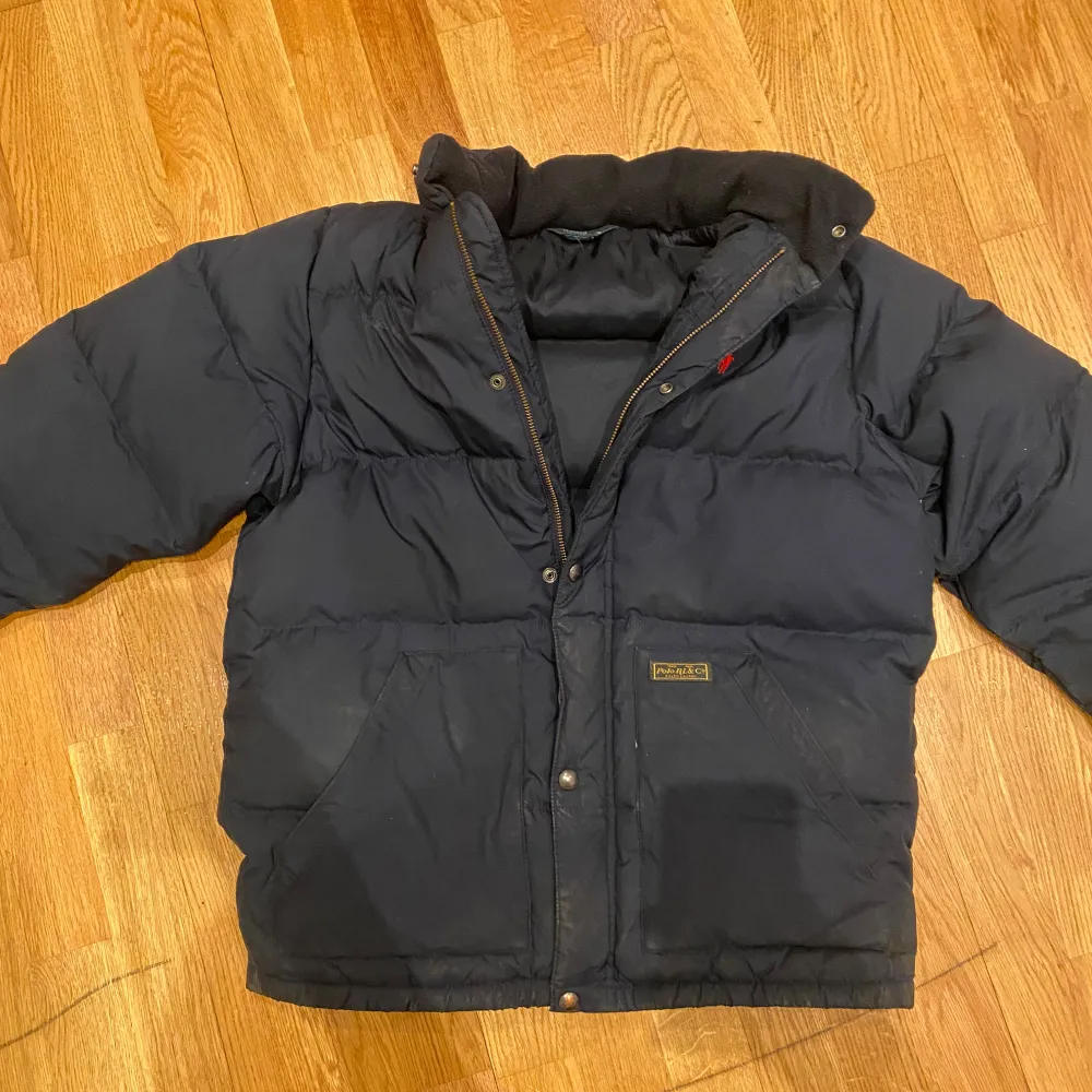 Säljer en mörkblå dunjacka från Polo Ralph Lauren med klassisk röd logga på bröstet. Jackan har hög krage, tryckknappar och dragkedja samt stora fickor framtill. Perfekt för kalla dagar och har en tidlös look.. Takit.