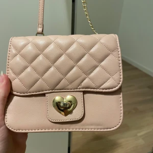 Beige quiltad axelväska från Glitter - Söt beige axelväska från Glitter med quiltat mönster och guldfärgad hjärtformad låsdetalj. Väskan har kedjerem och är perfekt för att bära det viktigaste. Stilren och trendig design som passar till många outfits.