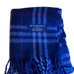 Blå rutig halsduk från Burberry - Säljer en snygg blå rutig halsduk från Burberry i 100% kashmir. Den har ett klassiskt rutmönster med inslag av rött och vitt, och fransar i ändarna. Perfekt för att hålla sig varm och stilren under kyliga dagar. 🧣