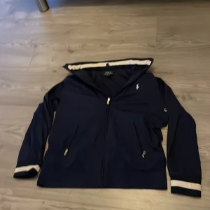 Marinblå jacka från Ralph Lauren - Säljer en stilren marinblå jacka från Ralph Lauren med dragkedja och broderad logga på bröstet. Jackan har långa ärmar och vita detaljer vid ärmslut och huva. Perfekt för en casual look!