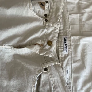 Vita byxor från Lois - Snygga vita byxor från Lois med hög midja och bootcut-stil. Byxorna har en dragkedja och knappar framtill samt fickor bak med Lois-logga. Perfekta för en stilren look.