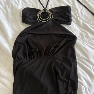 Svart topp från Sexy Silk - Snygg svart topp med halterneck och dekorativa metallringar framtill. Klänningen har en öppen rygg och är tillverkad i ett mjukt material som ger en elegant look. Perfekt för en kväll ute! Gissar på storlek XS