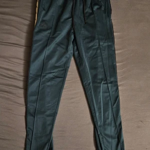 Mörkgröna träningsbyxor från Kappa med knappar - Säljer ett par mörkgröna träningsbyxor från Kappa med vita sidor och svarta Kappa-loggor längs benen. Byxorna har knappar längs hela sidorna och dragsko i midjan. Perfekta för en sportig look.