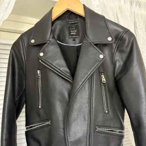 Svart skinnjacka med dragkedjor från Tri Outerwear - Säljer en svart skinnjacka från Tri Outerwear med klassisk bikerstil. Jackan har flera silvriga dragkedjor framtill och breda slag. Perfekt för dig som vill ha en tidlös och cool look.