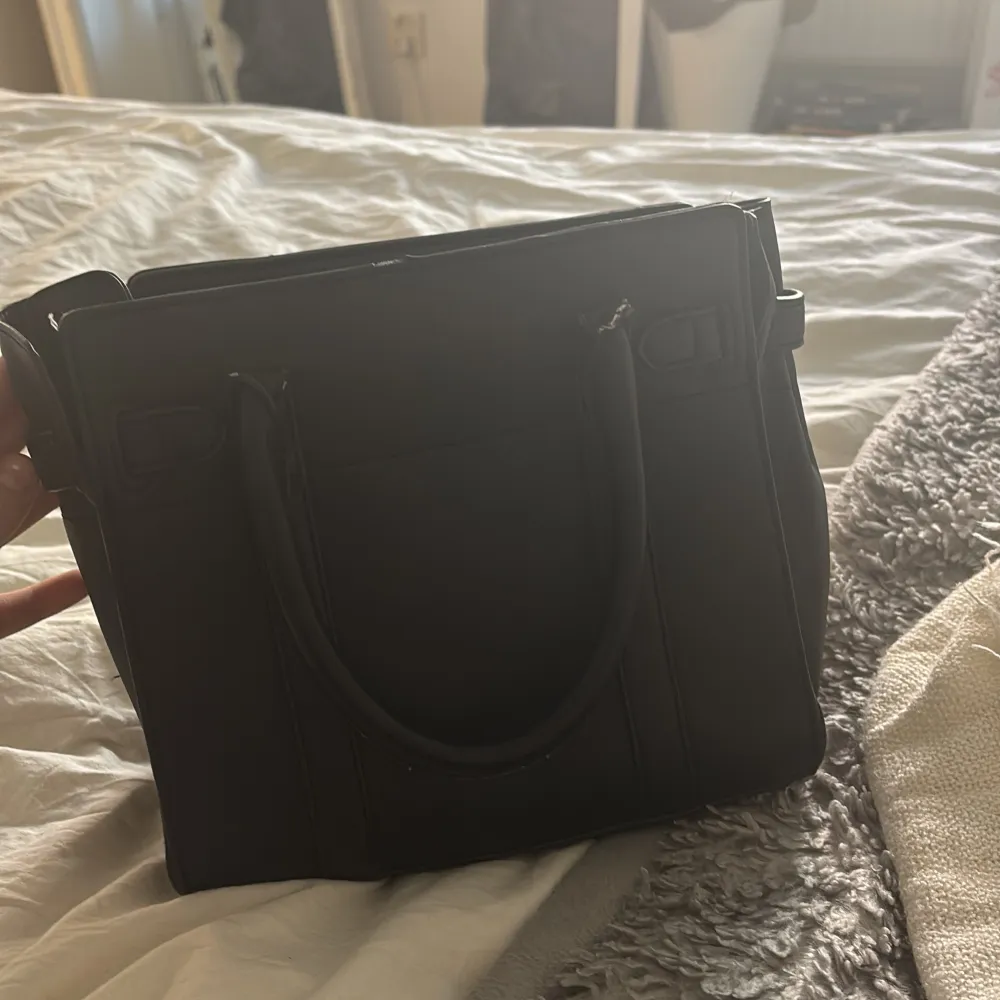 Säljer en stilren svart handväska med två handtag och snyggt guldfärgat spänne framtill. Väskan har flera fack inuti, inklusive ett med dragkedja. Perfekt för dig som vill ha en klassisk och rymlig accessoar till vardagen.. Laukut & Käsilaukut.