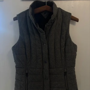 Grå quiltad väst från Tommy Hilfiger - Snygg grå quiltad väst i nyskick från Tommy Hilfiger med hög krage och dragkedja samt knappar framtill. Köpt på TH i Kuala Lumpur för 1400kr