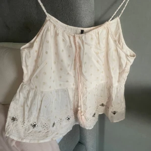 Ljusrosa linnetopp med guldprickar och broderi - Supersöt ljusrosa linnetopp med tunna axelband, guldprickar och broderade detaljer längst ner. Toppen har en volangkant och dekorativ snodd framtill. Perfekt för varma dagar!