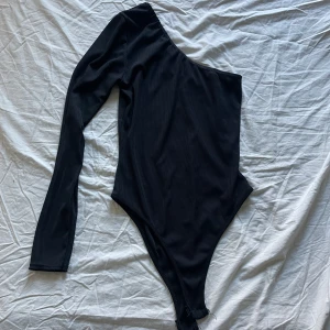 Svart oneshoulder bodysuit från Boohoo - Snygg svart oneshoulder bodysuit från Boohoo. Perfekt för en stilren look. Passar utmärkt till både jeans och kjol för en trendig outfit.